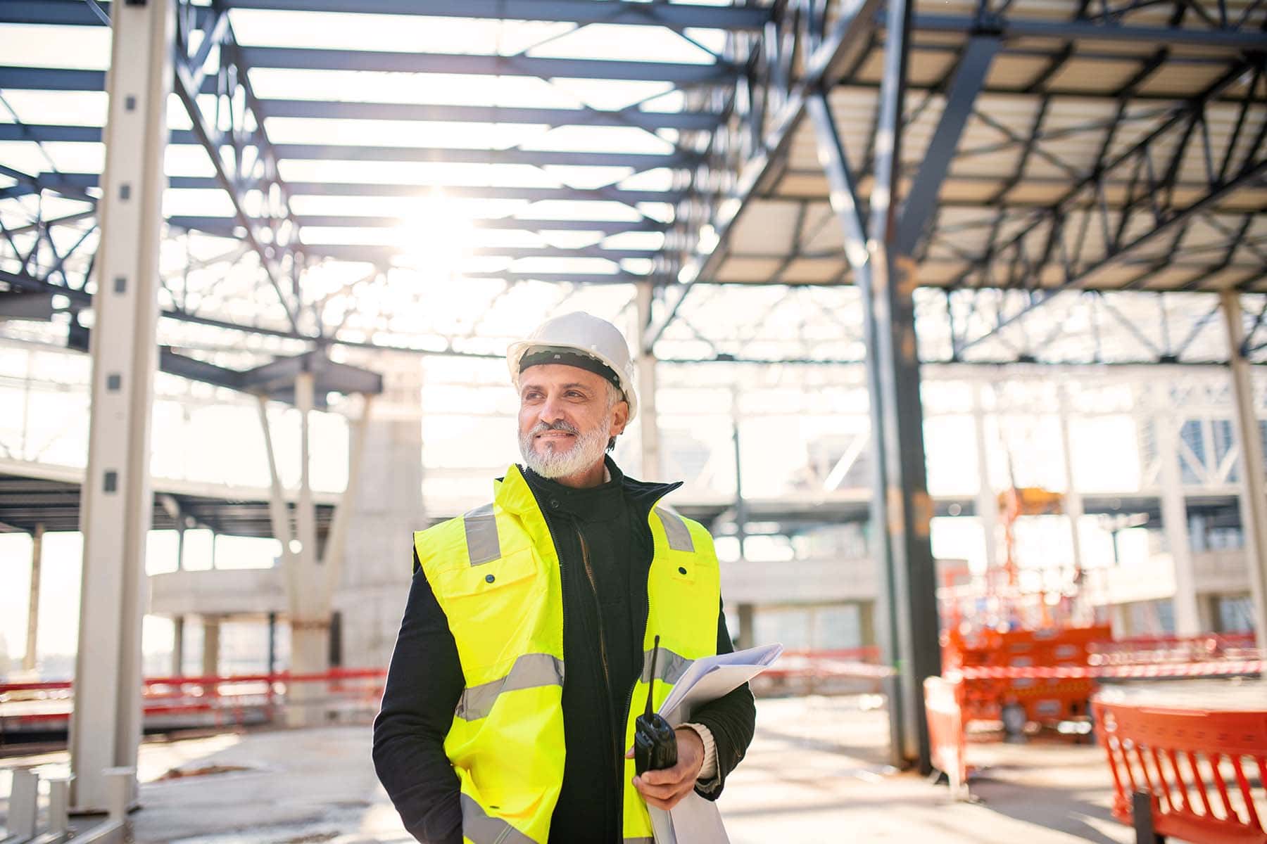 Bauingenieur mit Helm, Sicherheitsweste und Walkie-Talkie befindet sich auf einer Baustelle und blickt zufrieden in die Ferne.