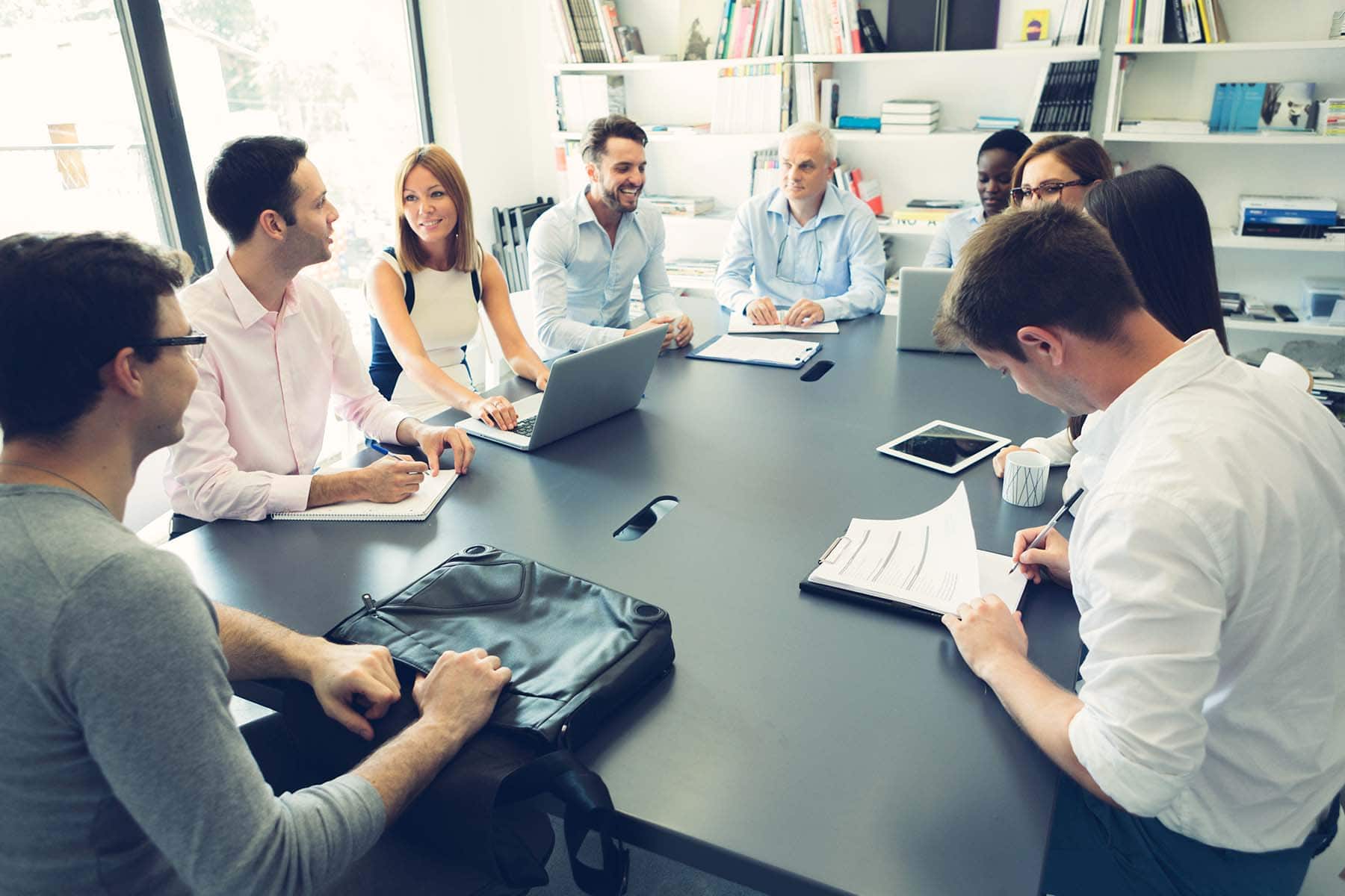 Eine Gruppe von Personen sitzt um einen großen Tisch, um ein Meeting abzuhalten. Alle haben entweder Laptop oder Stift und Papier vor sich, um das Reporting zu besprechen.