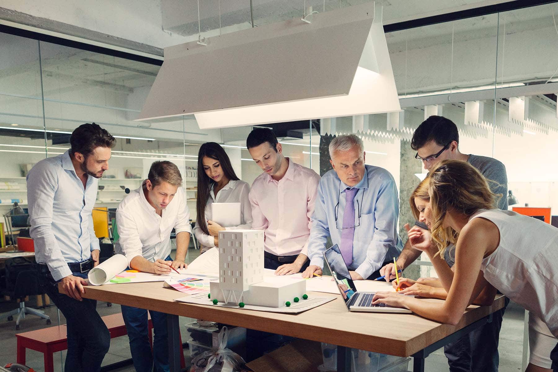 Gruppe von Personen steht um ein Architektur-Modell. Alle schauen sehr konzentriert. Mit einem Laptop sowie Stift und Papier werden wichtigen Informationen zur Ressourcenplanung festgehalten.